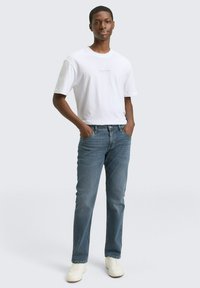 Weißes Baumwoll-T-Shirt mit kurzen Ärmeln, kombiniert mit hellblauen Jeans und weißen Sneakers. Das Modell steht mit den Händen in den Taschen vor einem neutralen Hintergrund.