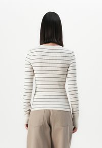 Vrouw met steil zwart haar, gekleed in een witte blouse met lange mouwen met dunne zwarte horizontale strepen en beige broek, van achteren gezien.