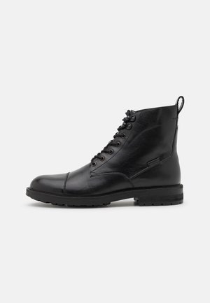 Bottine noire en cuir à lacets avec bout renforcé, semelle robuste et languette à l'arrière, vue de profil sur un fond uni.