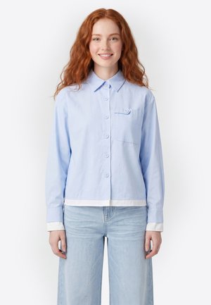 Junge Frau mit roten Haaren, die lächelt, trägt ein hellblaues Hemd mit Knöpfen und langen Ärmeln sowie hellblaue Jeans, steht vor einem weißen Hintergrund.