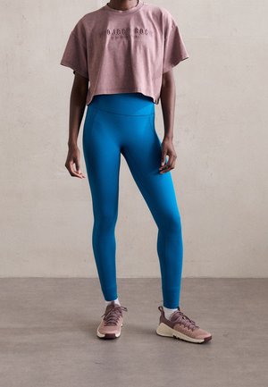 Personne portant un t-shirt court mauve avec le texte « Project Rock » et des leggings taille haute bleu vif, debout sur un sol gris contre un mur neutre.