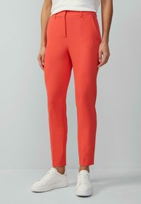 Helle, taillierte orangefarbene Hose mit schlanker Passform, gefertigt aus glattem Stoff. Kombiniert mit weißen Sneakers, die ein sauberes Design und minimale Details aufweisen.