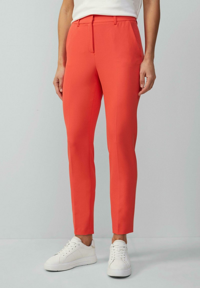 Helle, taillierte orangefarbene Hose mit schlanker Passform, gefertigt aus glattem Stoff. Kombiniert mit weißen Sneakers, die ein sauberes Design und minimale Details aufweisen.