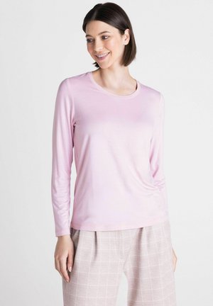Vrouw met kort donker haar die een lichtroze shirt met lange mouwen en lichtgekleurde geruite broek draagt, glimlachend en naar de zijkant kijkend.