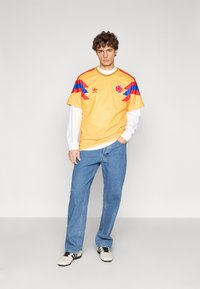 Maglia sportiva gialla con strisce diagonali blu e rosse, scollo rotondo, con logo ricamato. Abbinata a jeans in denim azzurro chiaro.