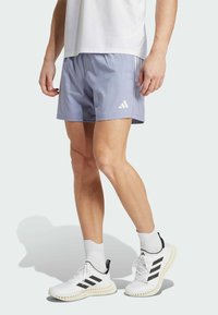 Pantalones cortos deportivos de color púrpura claro con una cintura elástica, que cuentan con tres franjas blancas y el logo de Adidas. Combinados con zapatillas blancas.