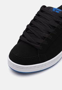 Baskets en daim noir avec lacets plats noirs, semelle intermédiaire blanche et détail de semelle extérieure bleu. Bout arrondi et perforations latérales pour la respirabilité.