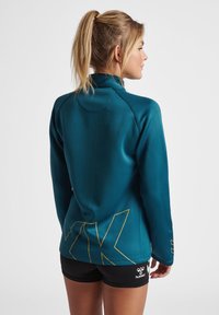 Turkusowa bluza sportowa z wysokim kołnierzem, długimi rękawami i żółtymi akcentami graficznymi na plecach, w połączeniu z czarnymi szortami. Gładka tekstura.