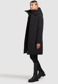 Long manteau noir avec un col montant, doté d'une fermeture éclair à l'avant, de poches latérales et d'un tissu lisse et résistant à l'eau. Associé à des bottes noires.