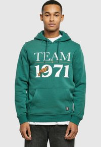 Zöld kapucnis pulóver, melyen fehér színnel "TEAM 1971" felirat található, kenguruzsebbel és egy kis címkével az alsó bal oldalon.