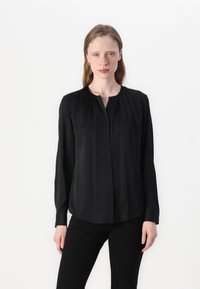 Blusa nera a maniche lunghe realizzata in tessuto leggero e liscio. Presenta un colletto plissé e una chiusura con bottoni. Abbinata a pantaloni neri.