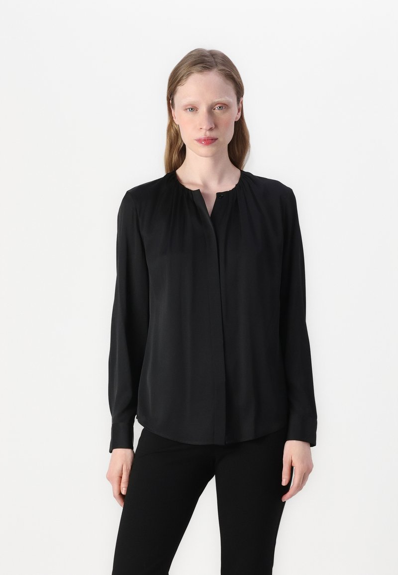 Blusa nera a maniche lunghe realizzata in tessuto leggero e liscio. Presenta un colletto plissé e una chiusura con bottoni. Abbinata a pantaloni neri.