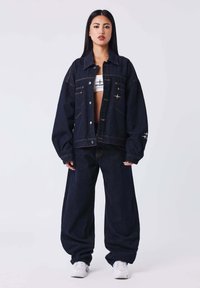 Chaqueta de mezclilla oscura de corte amplio, con bolsillos frontales y costuras en contraste, combinada con pantalones vaqueros oscuros de pierna ancha y zapatillas blancas.