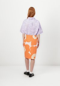 Chemise à manches courtes à motif floral violet et jupe crayon orange avec de grandes formes de fleurs blanches. Des chaussures noires et des chaussettes blanches sont portées.