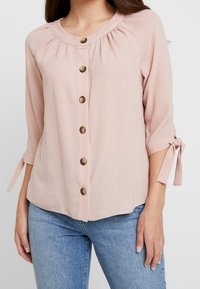 Blusa rosa chiaro con scollatura tonda, dettagli arricciati e sei bottoni marroni. Presenta maniche a tre quarti con fiocchi ai polsi.