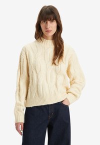 Pull en mailles torsadées crème à col haut, manches longues et ourlet à côtes, porté avec un denim sombre. Motif texturé sur le devant.