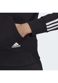 Main d'une personne dans la poche avant d'un sweat-shirt Adidas noir avec un motif trois bandes blanches sur la manche et le logo Adidas près de la poche.