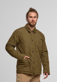 Quiltowana oliwkowa kurtka z kołnierzem z materiału sztruksowego, przednią kieszenią i zapięciem na zatrzaski; charakteryzuje się teksturowaną powierzchnią i luźnym krojem.