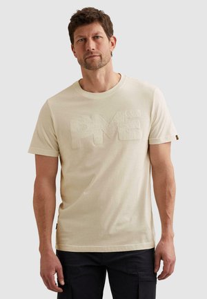 Muž stojaci pred jednofarebným pozadím, oblečený v béžovom tričku s krátkym rukávom s vystúpeným nápisom "PME" a čiernych nohaviciach.