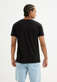 Camiseta de algodón negra de manga corta, con cuello redondo y un corte relajado. El modelo lleva puestos unos pantalones cortos de mezclilla azul claro. La vista desde atrás muestra una textura suave.