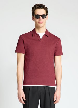 Uomo con occhiali da sole neri, camicia a maniche corte rosso scuro con colletto sopra una canottiera bianca e pantaloni neri, in piedi davanti a uno sfondo semplice.