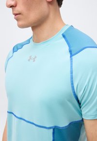 Άνδρας που φοράει αθλητική μπλούζα Under Armour σε ανοιχτό γαλάζιο με πιο σκούρες μπλε λεπτομέρειες και ραφές, στραμμένος προς τα αριστερά πάνω σε λευκό φόντο.