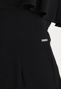 Czarna odzież z falbankowym dekoltem i metalowym detalem z logo. Tkanina ma gładką teksturę i dopasowany krój.