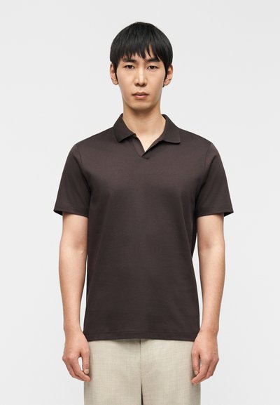 Jeune homme debout, face à l'avant, portant un polo à manches courtes marron foncé et un pantalon beige clair sur un fond uni.