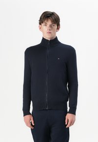 Tume sinine zip-up sviiter kõrge kaelusega, soonikute ja äärtega. Valmistatud pehmest kangast, millel on rindkere küljes väike logo.