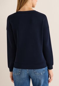 Marinefarbener Sweatshirt mit langen Ärmeln, weitem Ausschnitt und geripptem Saum. Weiches Material und lockere Passform. Kombiniert mit hellblauen Jeans.