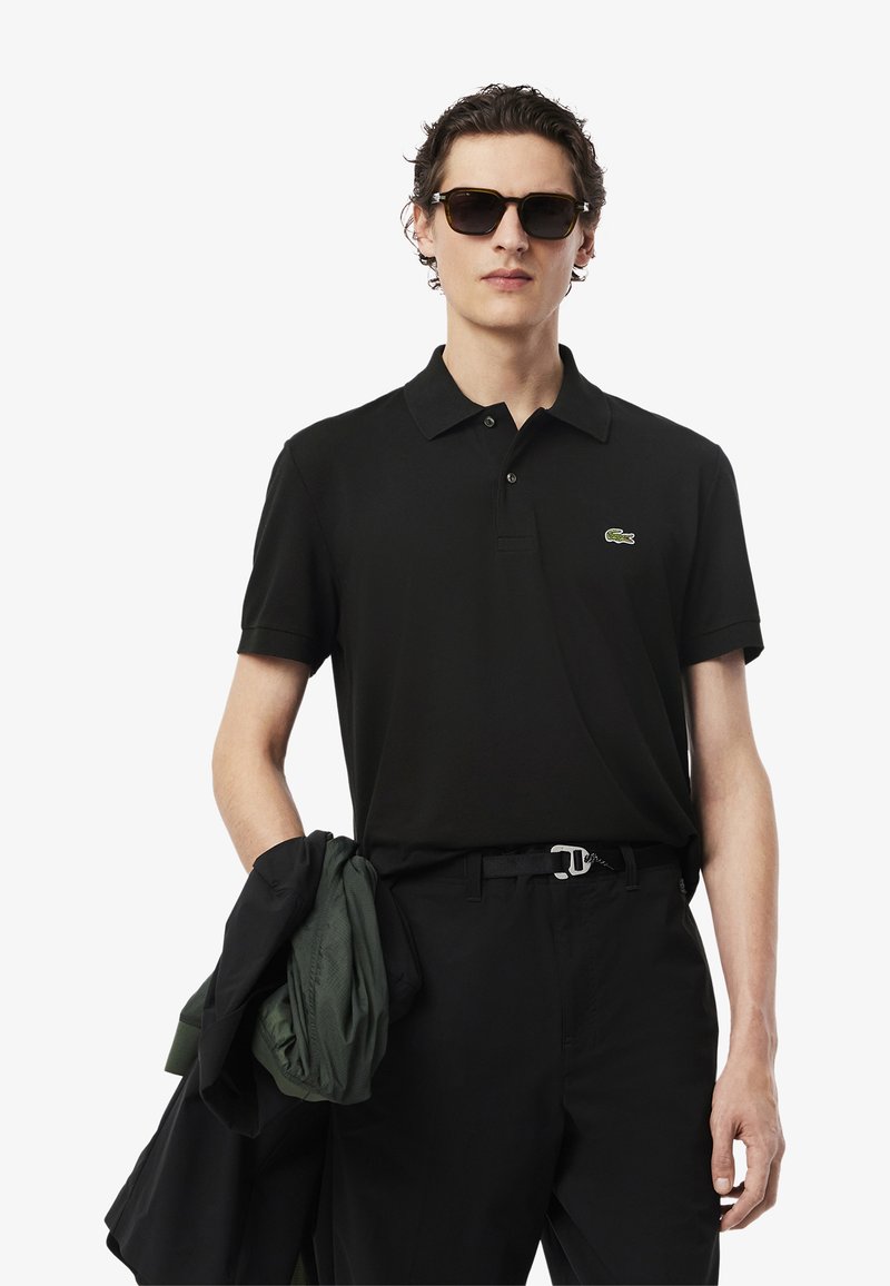 Camisa polo negra con cuello y logo verde bordado, combinada con pantalones negros. Sostiene una chaqueta verde sobre un brazo y lleva gafas de sol.