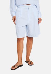 Lys blå linshorts med tilpasset liv, folder foran og en avslappet passform, parret med svarte flip-flops.