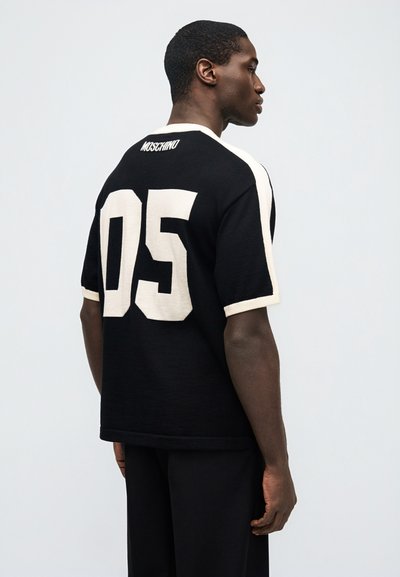 Άνδρας με μαύρο και λευκό πουκάμισο Moschino με αριθμό 05 στην πλάτη, στέκεται πλάγια μπροστά σε απλό ανοιχτό φόντο.