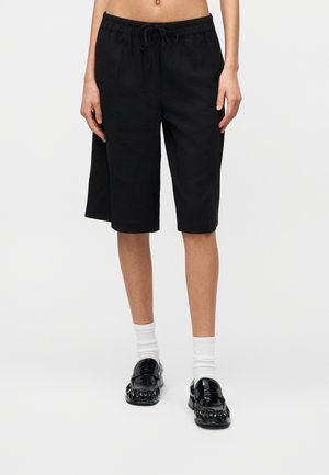 Personne portant un short ample noir mi-cuisse, des chaussettes blanches et des mocassins noirs brillants, vue de la taille aux pieds sur fond blanc.