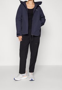 Veste imperméable marine avec capuche, haut ajusté noir, pantalon noir avec poches zippées, et chaussures de course grises avec des accents bleus et oranges.