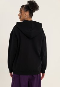 Personne aux cheveux bouclés coiffés en chignon haut avec une boucle d'oreille, portant un sweat à capuche noir et un pantalon violet, vue de profil sur un fond uni.