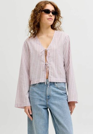 Femeie purtând ochelari de soare negri, o bluză largă în dungi cu șnururi în față și blugi albaștri deschiși cu talie înaltă, stând în picioare pe un fundal simplu.