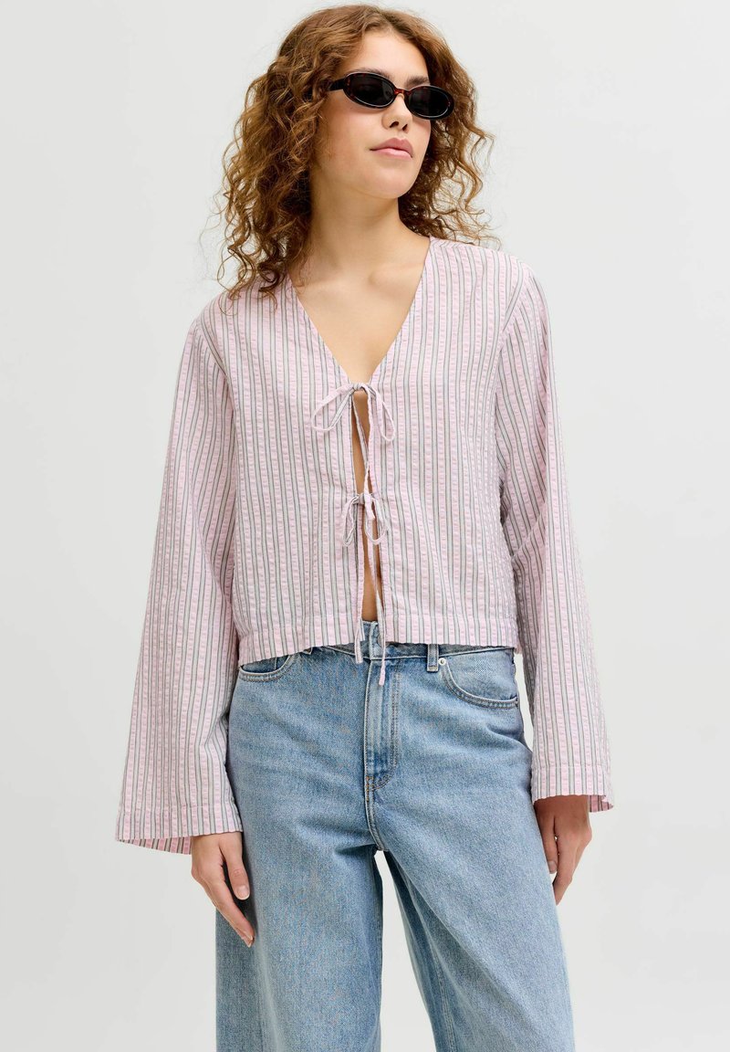 Kvinde iført sorte solbriller, løs, stribet bluse med bindebånd foran og lyseblå jeans med høj talje, stående foran en ensfarvet baggrund.