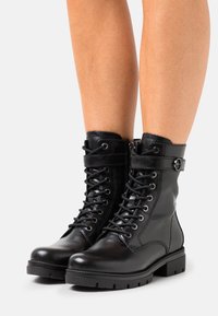 Bottines en cuir noir avec un laçage à l'avant, fermeture éclair sur le côté et détail de sangle avec boucle. Semelle en caoutchouc épaisse avec un motif texturé pour une adhérence optimale.