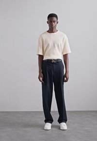 Cream ribbed short-sleeve shirt, black tailored trousers with side pockets, and white sneakers. Notable silver belt buckle with "M" detail.