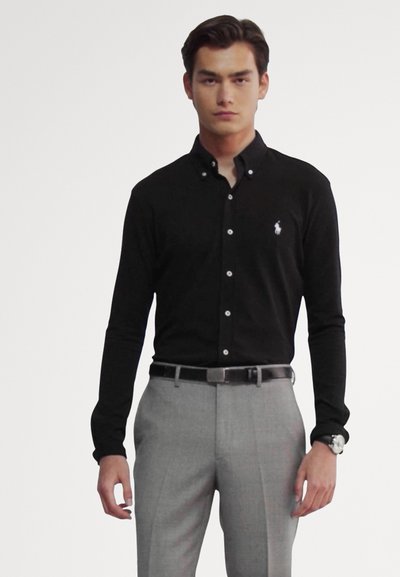 Zwarte langemouwen shirt met een button-down kraag, geborduurd logo op de borst, gecombineerd met lichtgrijze op maat gemaakte broek en een horloge.