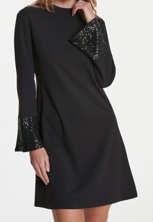 Femme portant une robe noire à manches longues avec de larges poignets pailletés, posant avec une main près du cou et l'autre le long du corps.