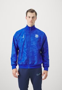 Blauwe zip-up jack van synthetisch materiaal, met een gestructureerd patroon, hoge kraag en Barça-logo op de linkerborst.