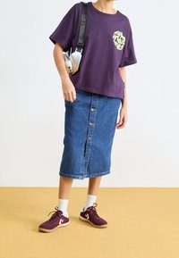 Person som bär en lila grafisk T-shirt, en knälång knappad jeanskjol, vita strumpor och lila sneakers, med en silverfärgad axelväska.