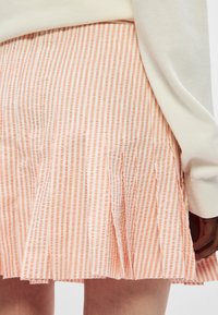 Plissé kort rokje in licht perzik met witte verticale strepen, met een textuurstof en een gladde tailleband, gedeeltelijk bedekt door een lichte top.