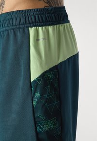 Heren sportshorts in donkerteal met een lichtgroen mesh paneel. Kenmerken geometrische groene patronen, elastische tailleband en DRYCELL branding.