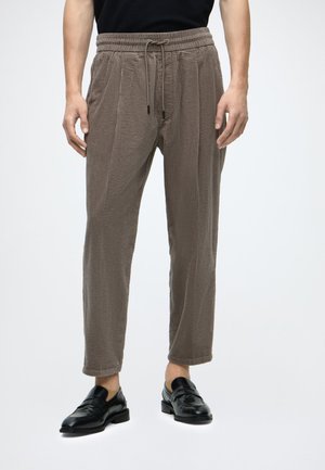 Man wearing brown textured drawstring pants and black leather loafers, standing against a plain light background.