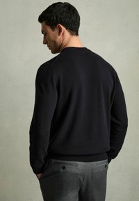 Marinblå tröja med ribbade ärmslut, bärs över ljusgrå byxor med bakfickor med knappstängningar, visas bakifrån.