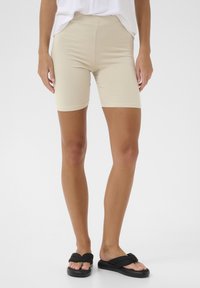 Pantalones cortos de bicicleta de mezcla de algodón beige con una cinturilla elástica, de longitud media hasta el muslo y textura suave, combinados con sandalias negras de fácil puesta.