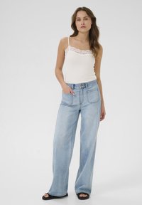 Top blanc sans manches avec bordure en dentelle, associé à un jean large taille haute bleu clair avec poches avant et arrière, et des sandales plates noires.
